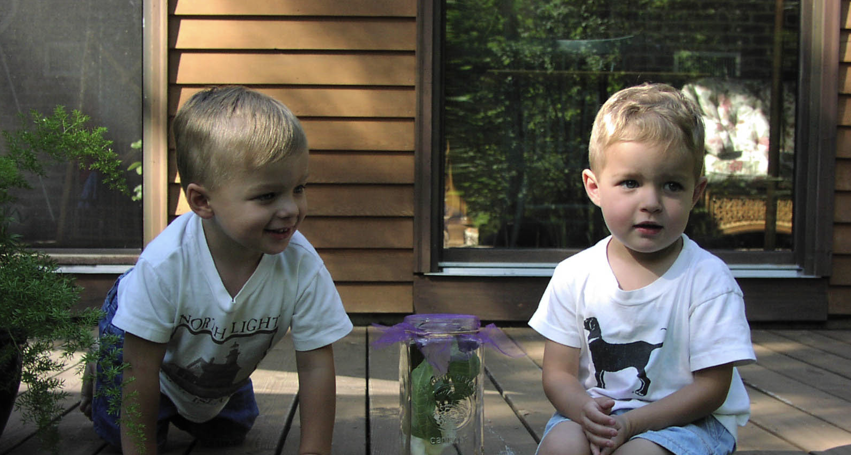 two toddlers sitting outside, between them is a jar with a flower in it