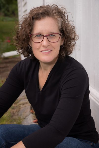 Professional author headshot of Loree Griffin Burns, Loree in a black v-neck sweater, blue jeans and red glasses looking at the camera